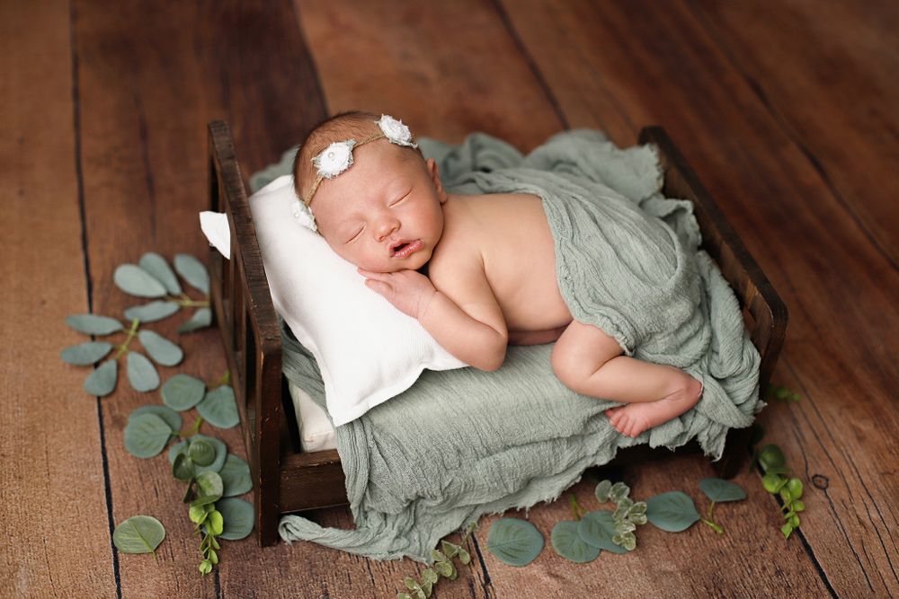 Sleeping newborn wrapped in soft sage green fabric on wooden floor with decorative eucalyptus leaves.