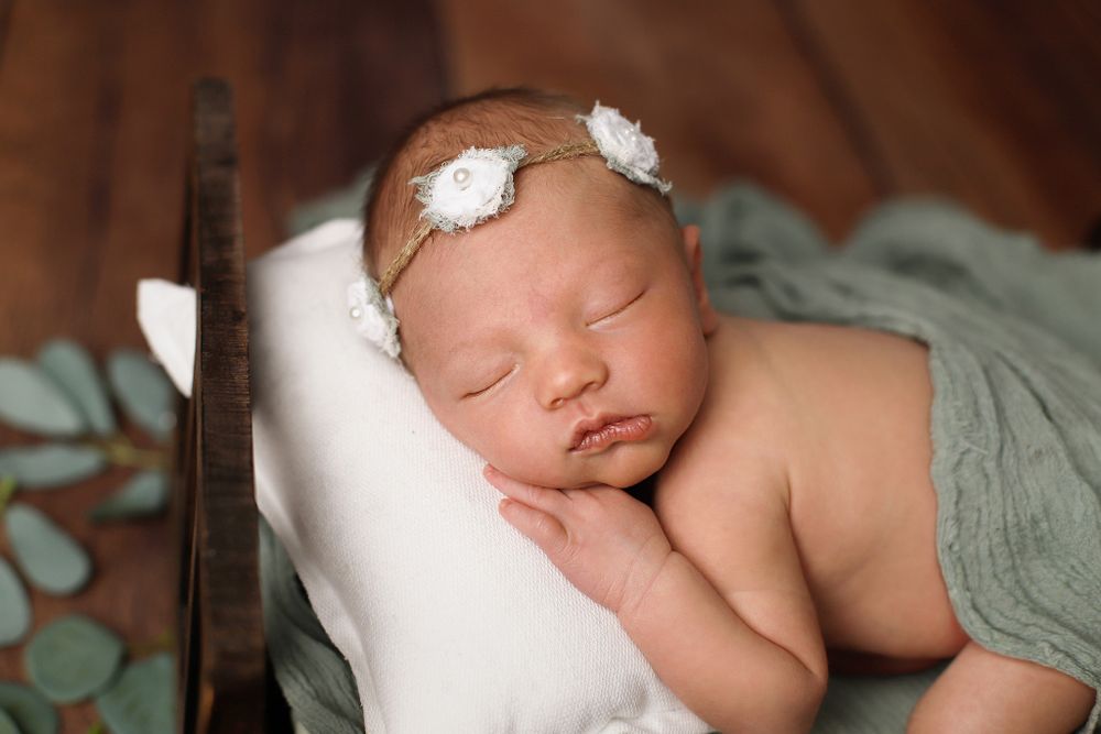 Peaceful sleeping newborn baby wearing delicate white floral headband wrapped in sage green fabric on white pillow.