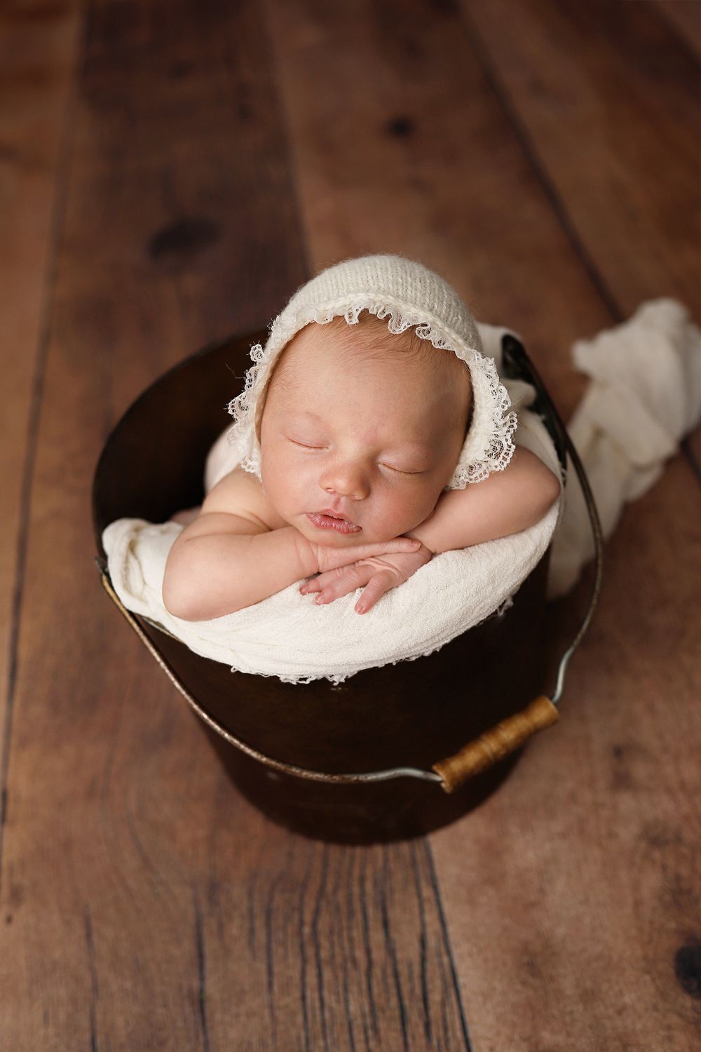 Sleeping newborn wearing delicate white bonnet nestles peacefully in dark wooden bucket on hardwood floor.