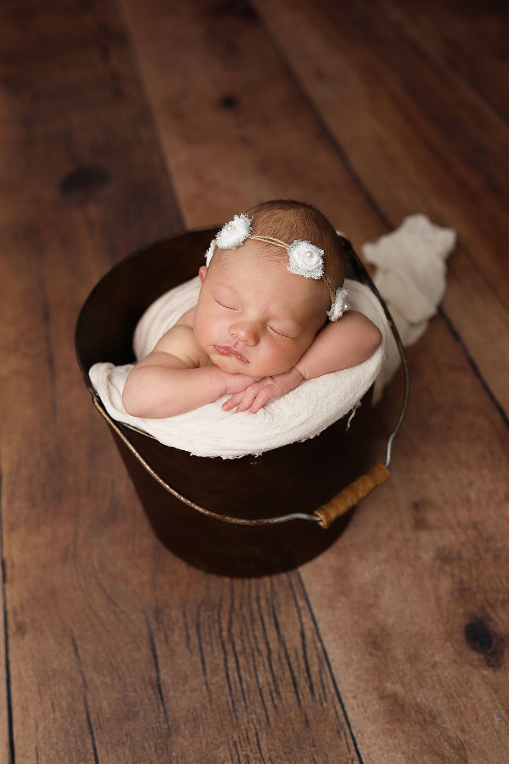 Sleeping newborn wearing delicate white floral headband rests peacefully in dark wooden bucket on rustic floor.