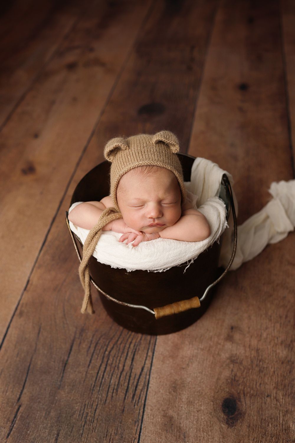 A sleeping newborn wearing a knit bear hat rests peacefully in a round basket on wooden flooring.