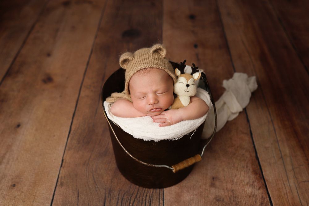 Sleeping newborn wearing bear hat lying in dark basket with stuffed animal on wooden floor.