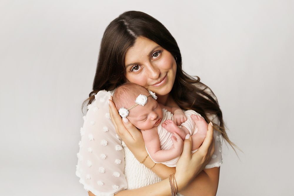Woman in white sweater holds sleeping baby against light background during newborn photo session.