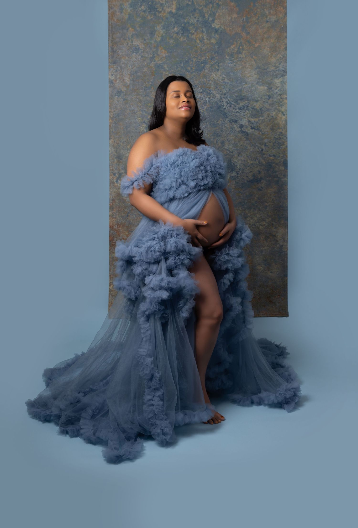 A maternity photo featuring an ethereal blue tulle gown with ruffled details against a textured backdrop.