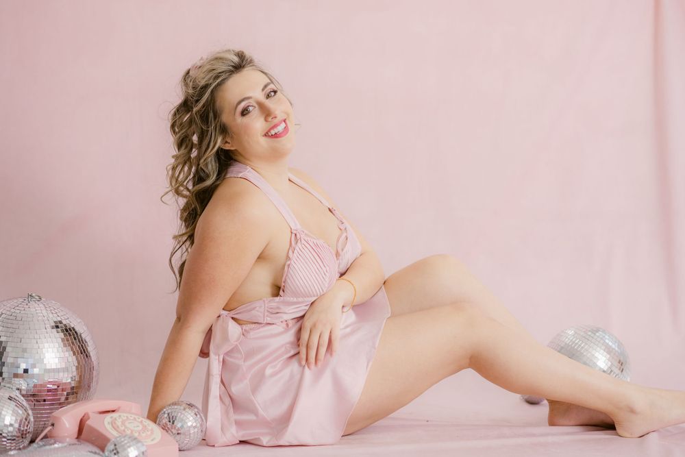 Person in pink silk dress poses on pink backdrop with silver disco balls for glamour boudoir photography session.