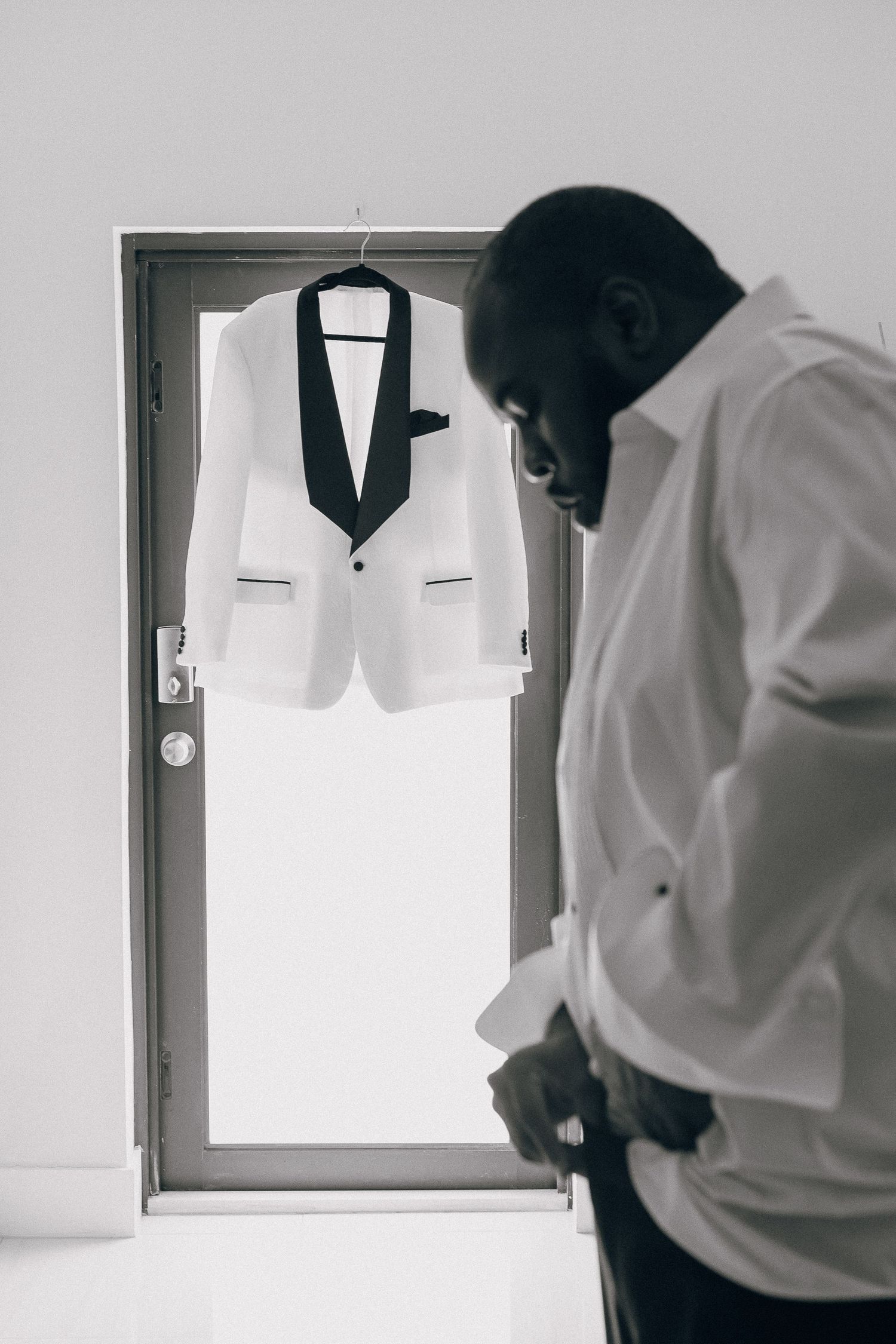 White tuxedo jacket displayed on hanger in doorway with someone preparing in foreground.
