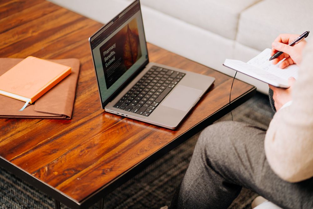 Laptop on wooden desk with orange notebook and someone taking notes.