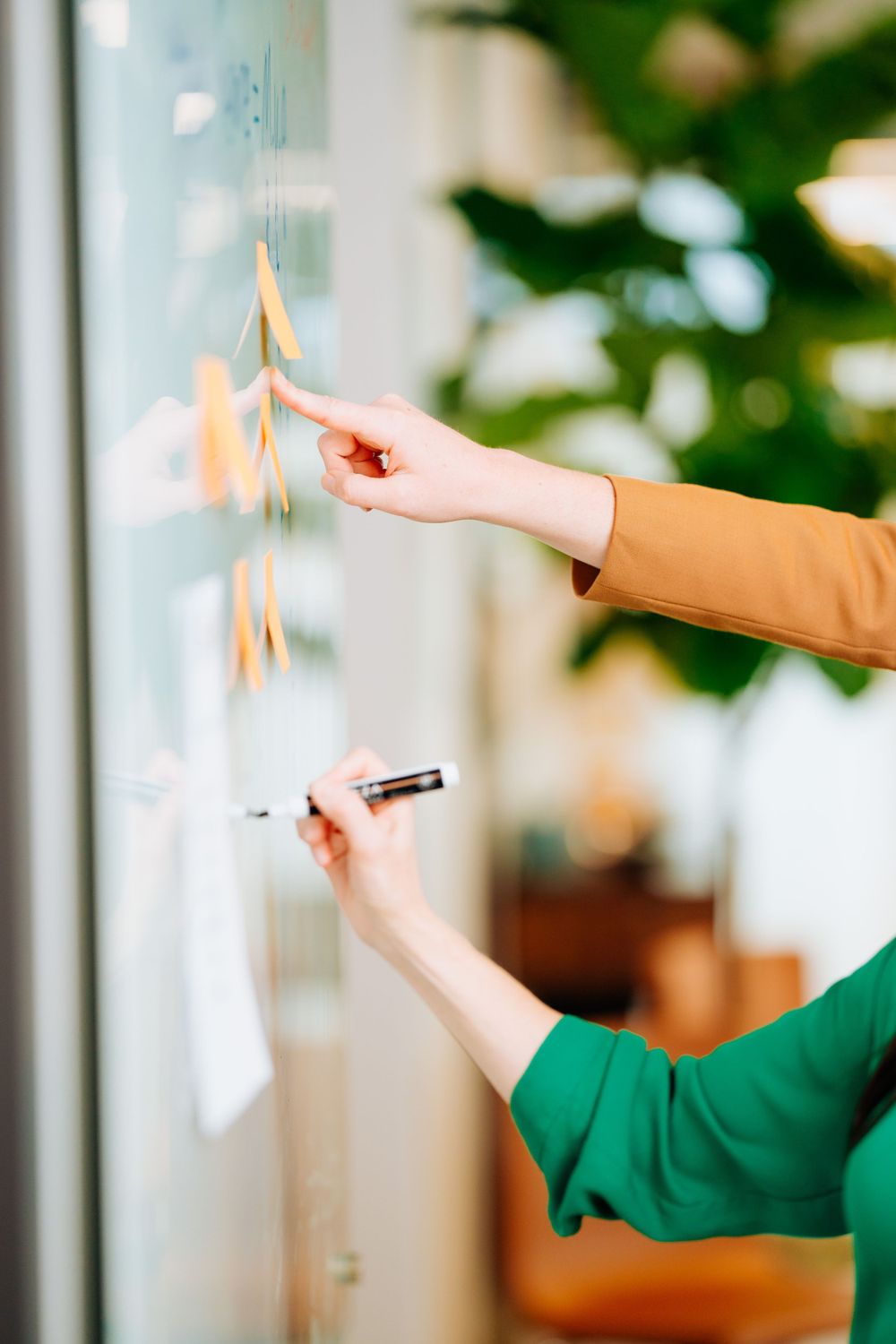 Hands placing sticky notes on a glass wall with plants in the background.