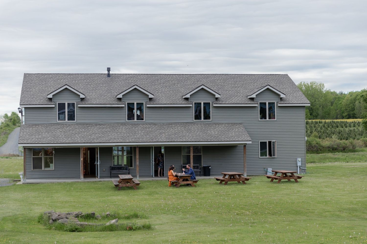 Large gray residential building with dormers and picnic tables on the lawn against an overcast sky.