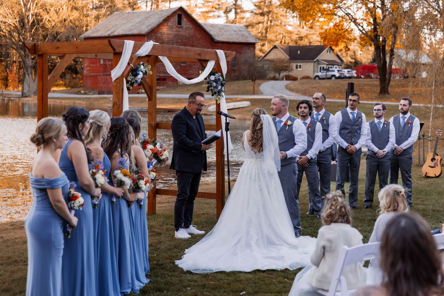 An outdoor wedding ceremony at sunset with bridesmaids in blue dresses and groomsmen in vests standing before a rustic barn.