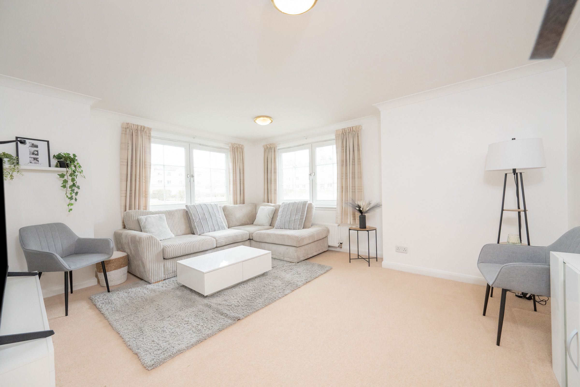 Modern bright living room with beige L-shaped sofa, grey chairs and white ottoman on grey rug in minimalist style.