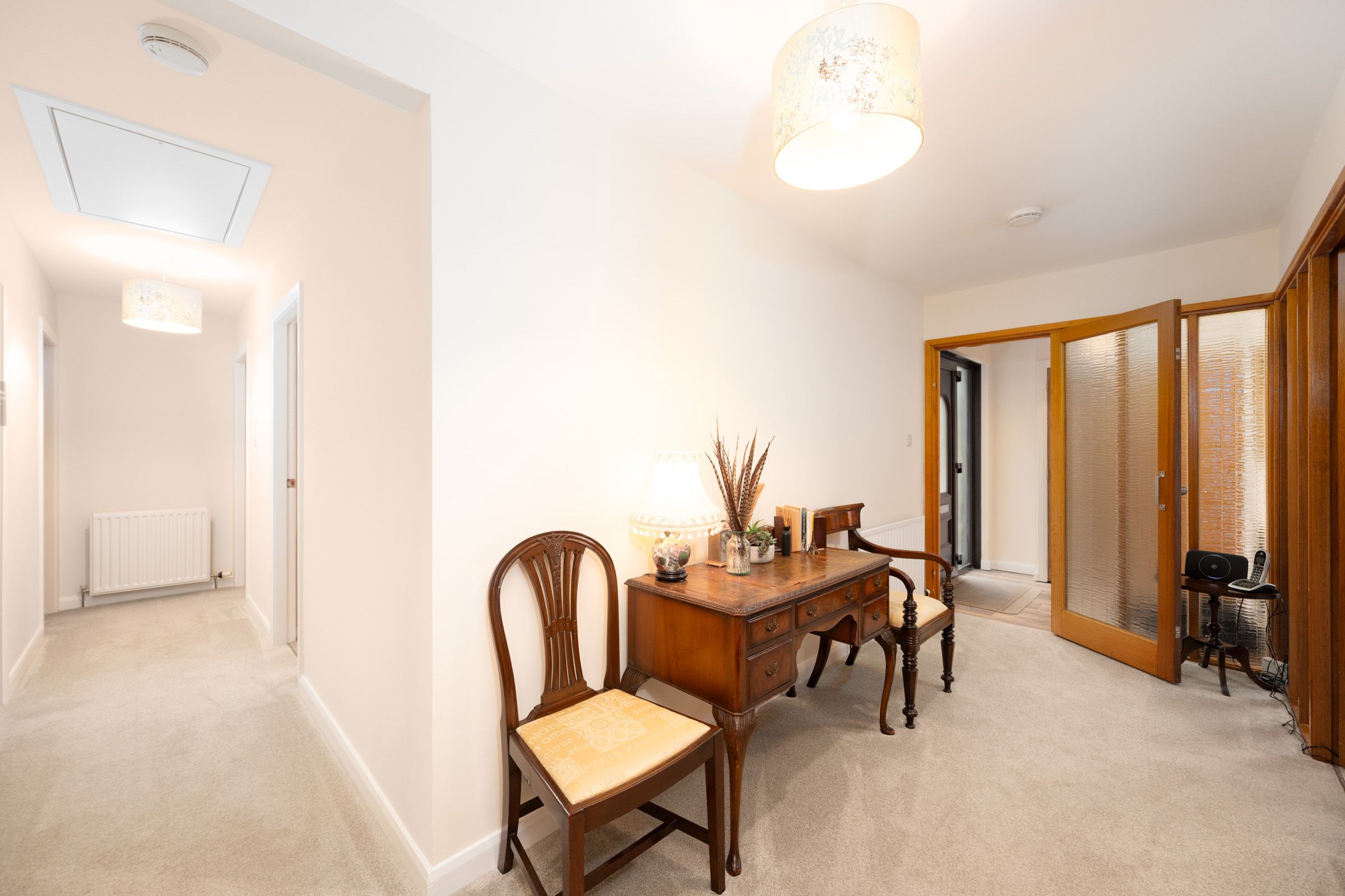 Interior hallway with beige carpet leading to a wooden desk and chair set with glass doors and white walls.
