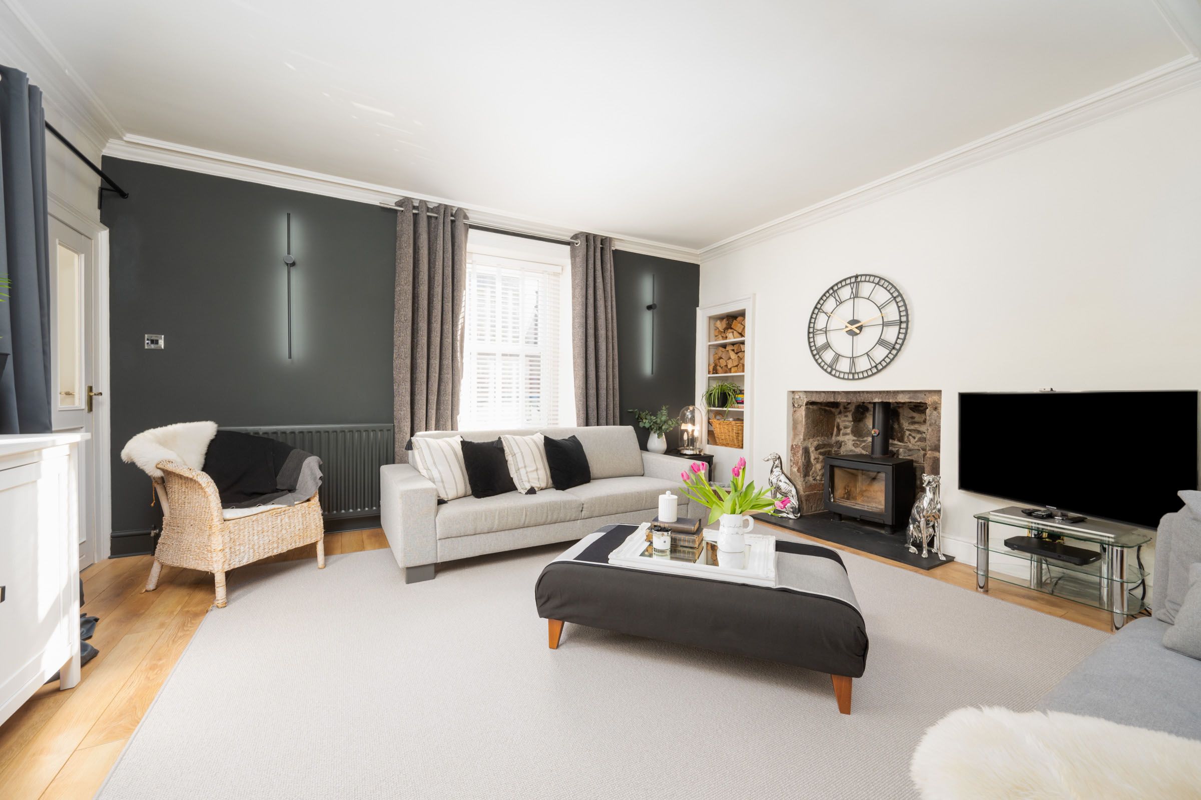 Modern living room with grey accent wall, beige sofa, dark ottoman, and decorative clock above fireplace.