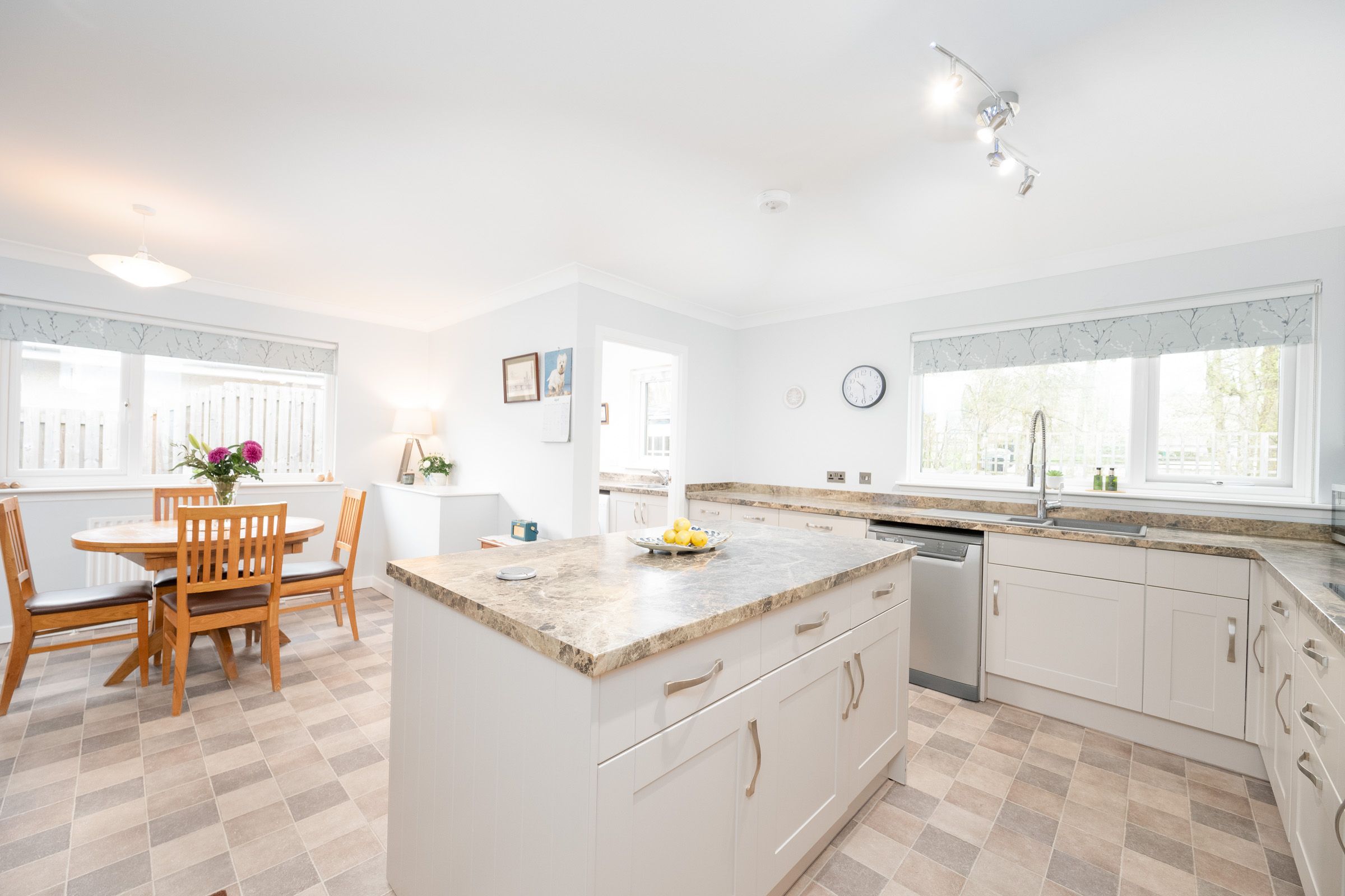 Modern white kitchen with granite island, checkered floor, and wooden dining set in bright sunlit space.