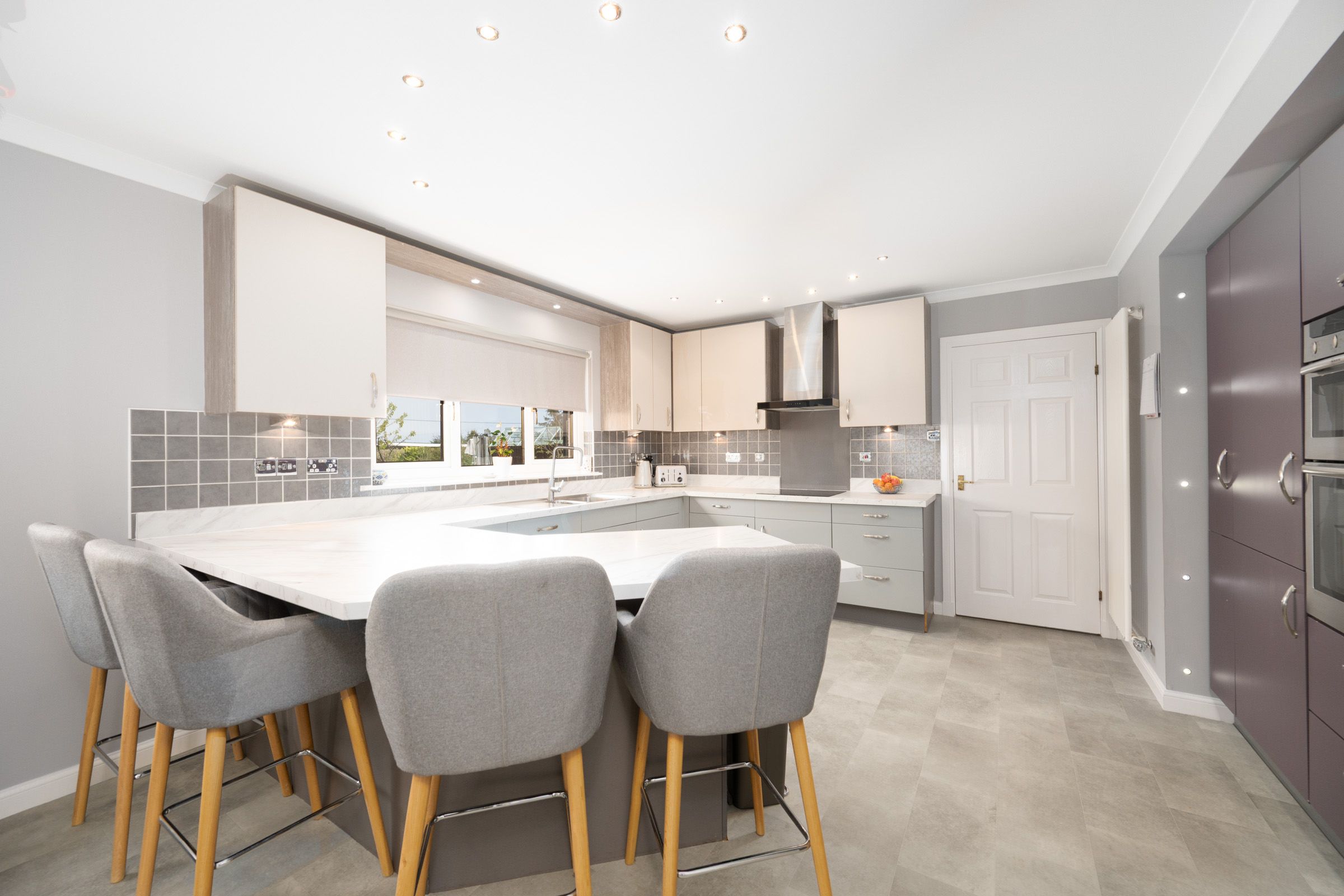 Modern white kitchen with gray upholstered bar stools features clean lines, light cabinetry and peninsula dining space.