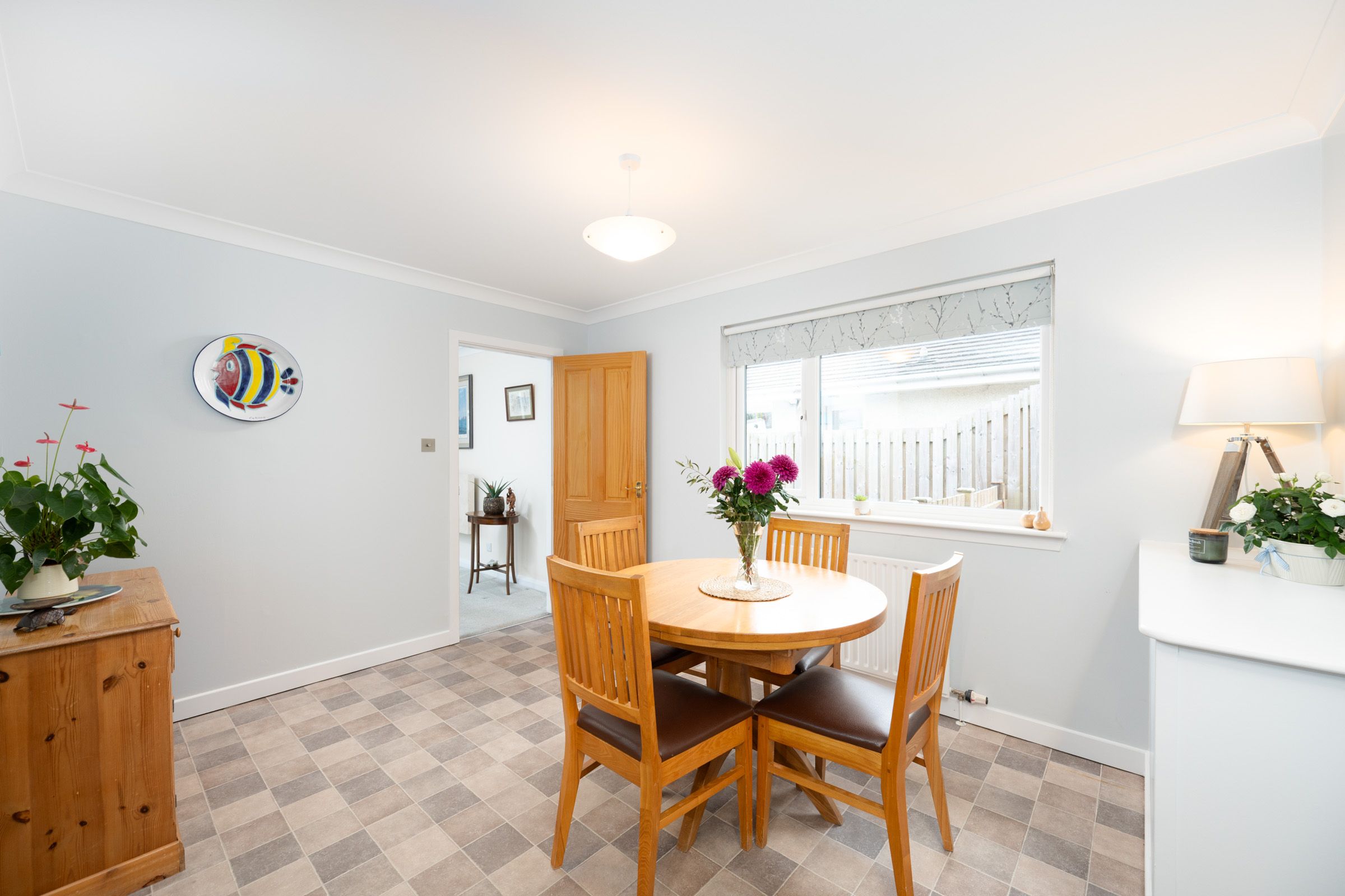 A bright dining room with wooden furniture, checkerboard flooring, and decorative plants by a window.