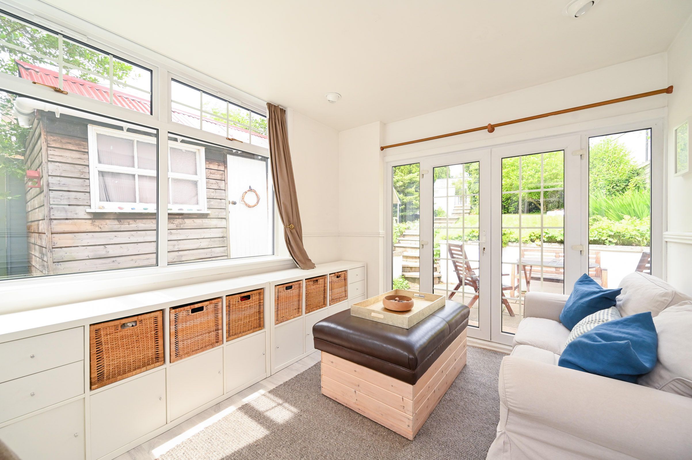 Bright sunroom with built-in storage benches, wicker baskets, and blue throw pillows features large windows overlooking greenery.