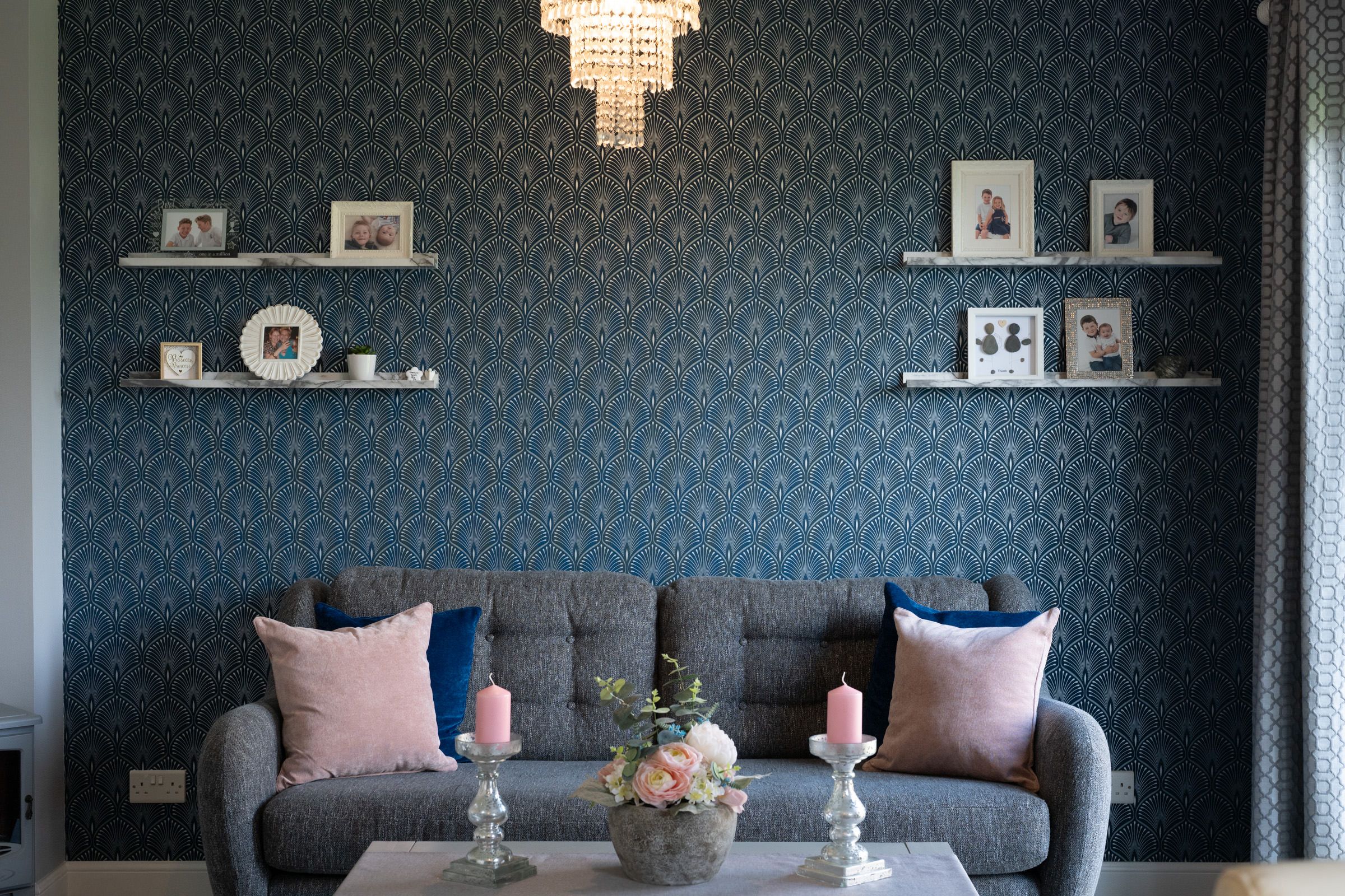 Modern living room with gray sofa against dark patterned wallpaper and floating shelves displaying framed photos.