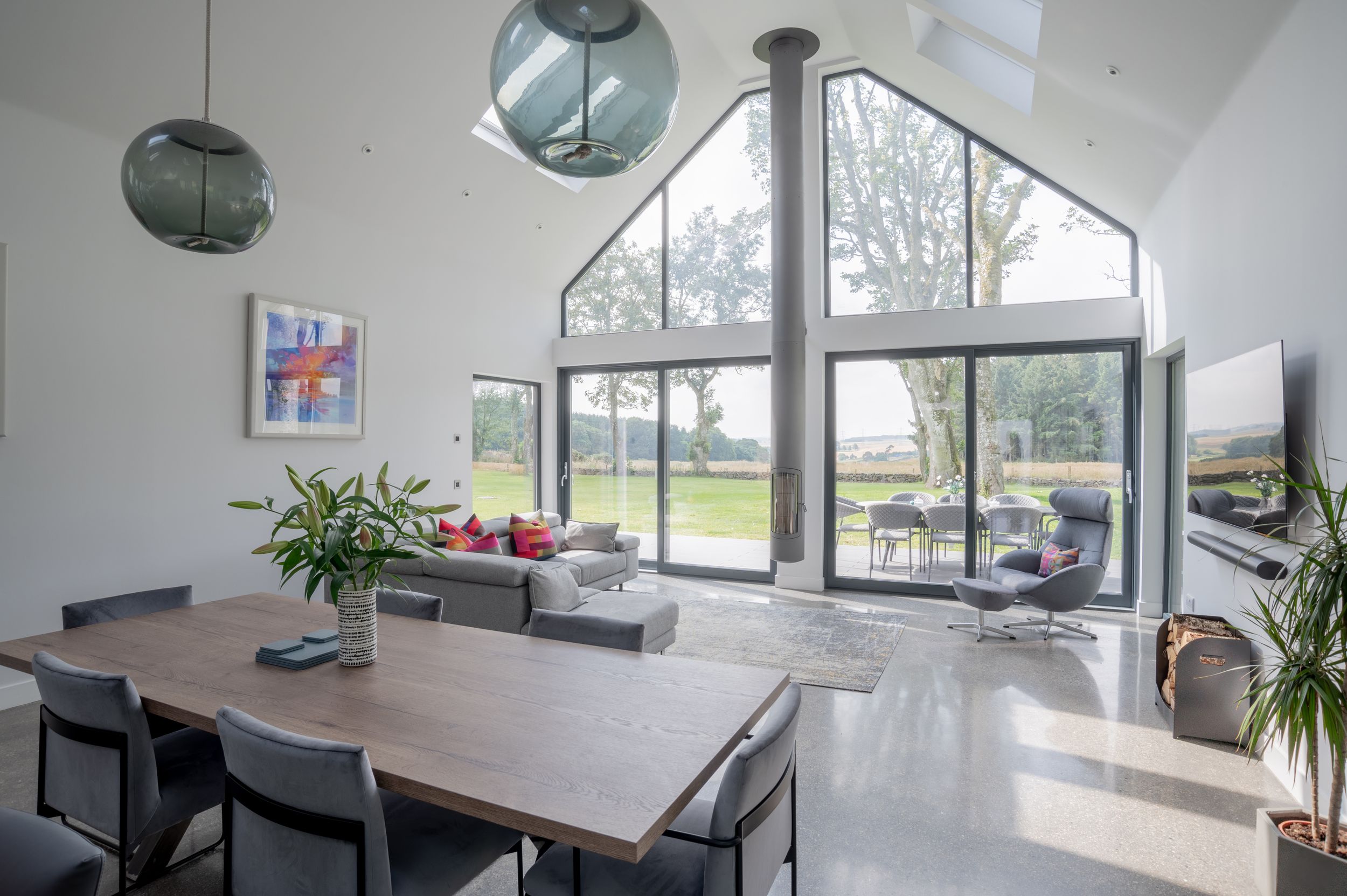 Modern dining room with large triangular window wall, wooden table, and pendant lights in a bright sunlit space.