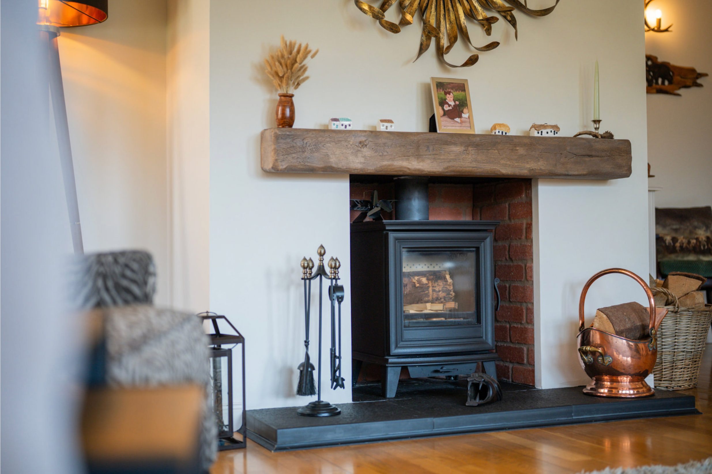 Cozy wood burning stove with rustic wooden mantel, fireplace tools and copper kettle in a traditional home interior.
