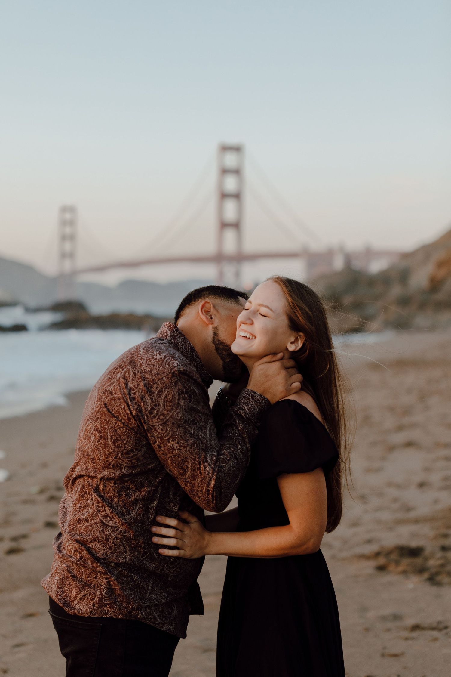 A romantic couple embraces on the beach with the iconic Golden Gate Bridge visible in the background at sunset.