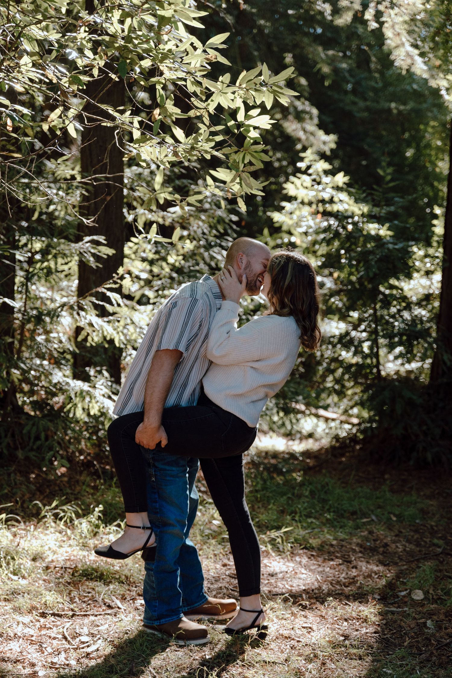 A romantic couple sharing an intimate moment while standing beneath sunlit trees in a wooded setting.