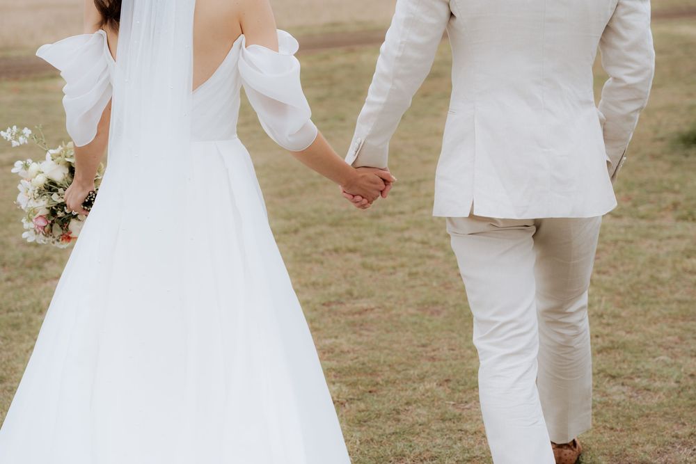 A bride in a white gown and groom in white suit hold hands while walking on grass during their wedding day.