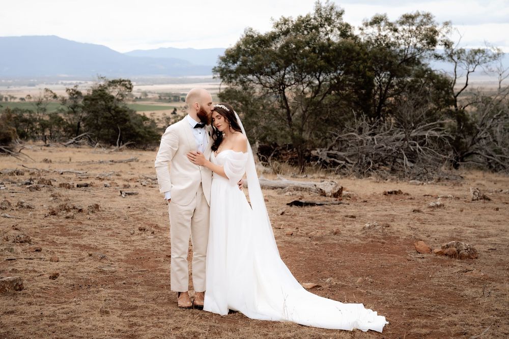 A couple in wedding attire share an intimate moment on a hillside overlooking a scenic desert landscape during sunset.