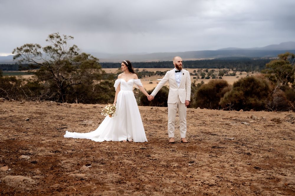 A wedding couple holds hands on a hilltop overlooking scenic mountains and valleys on a cloudy day.