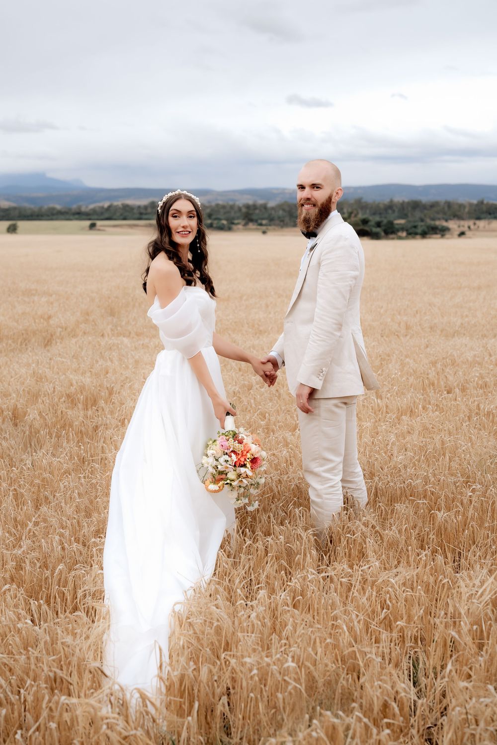 A wedding couple stands together in a golden wheat field with mountains in the background on an overcast day.