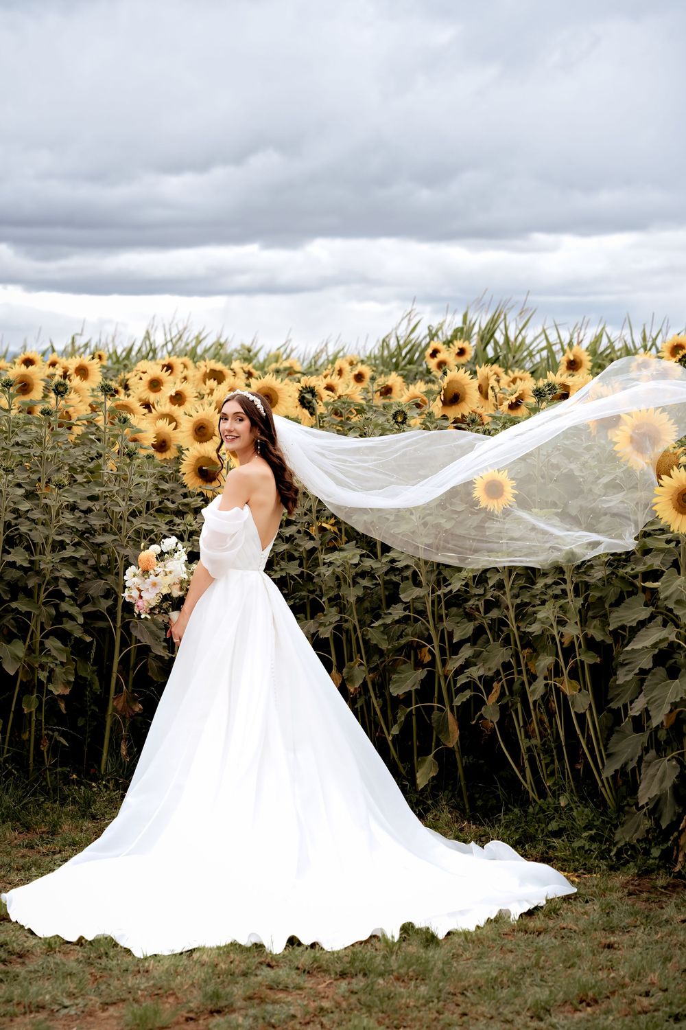 A flowy white wedding dress and flowing veil catch the breeze in front of a stunning field of bright yellow sunflowers.