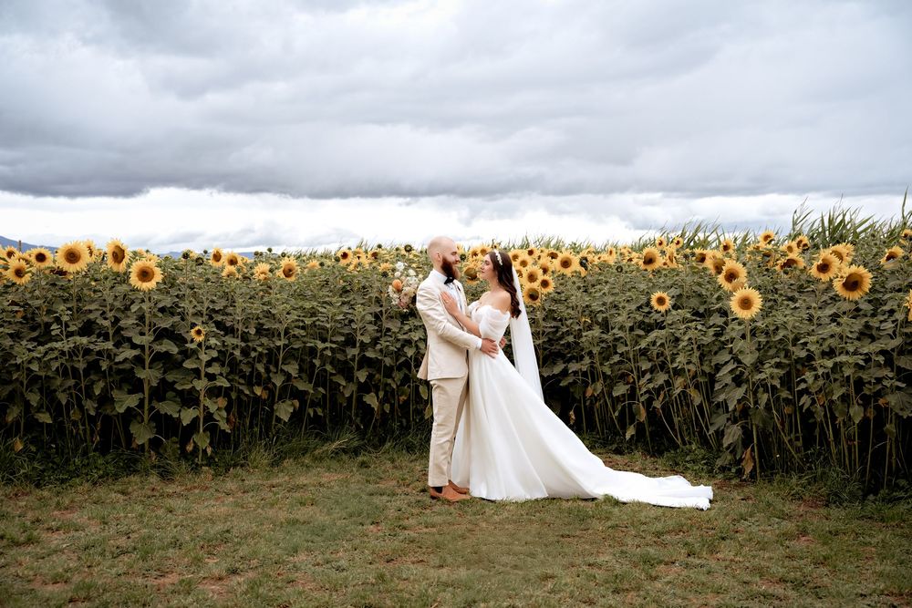 A couple stands intimately together in a sunflower field under dramatic cloudy skies during their wedding photoshoot.