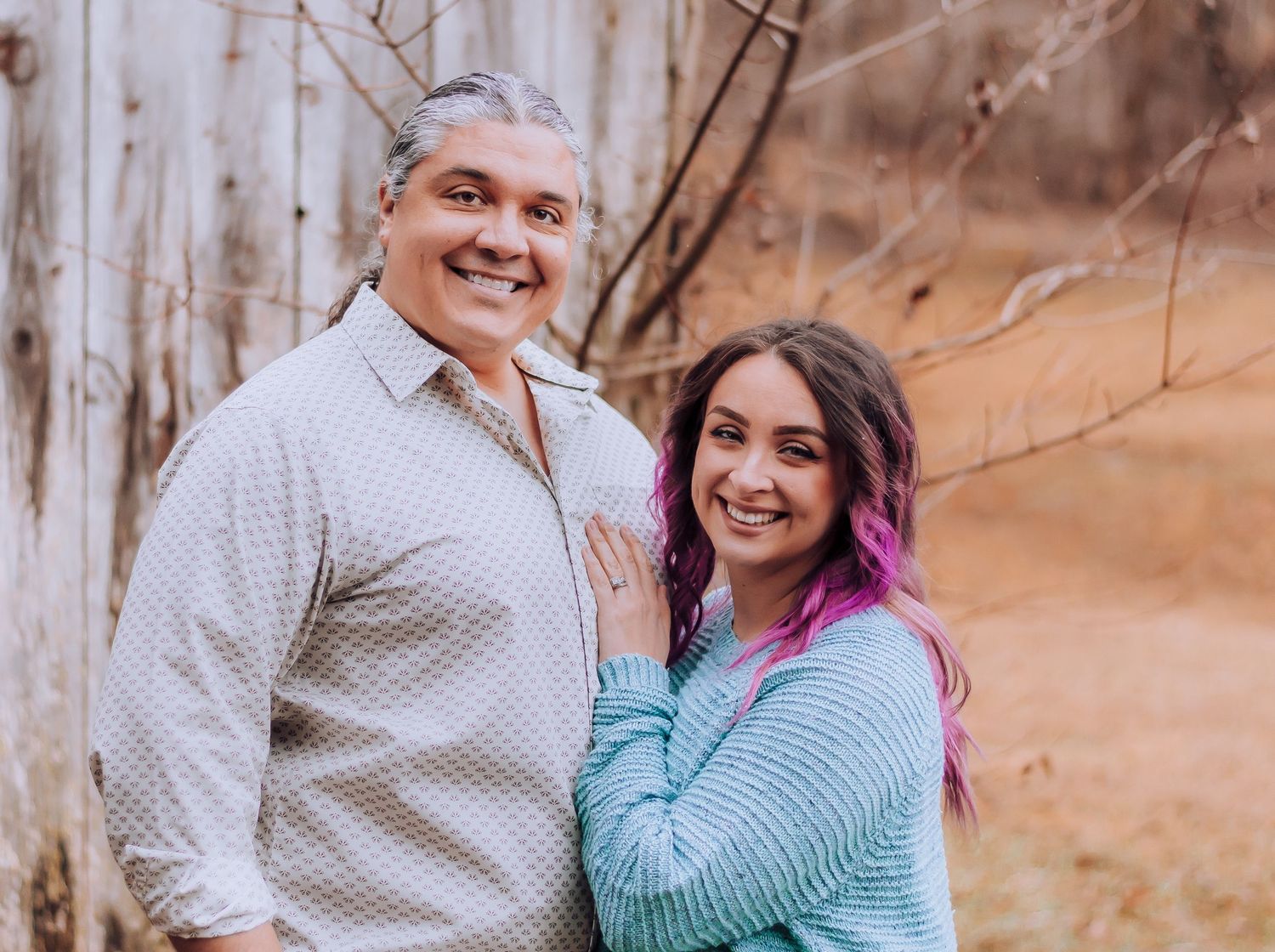 Two people embrace and smile outdoors during autumn with a wooden fence and brown background.