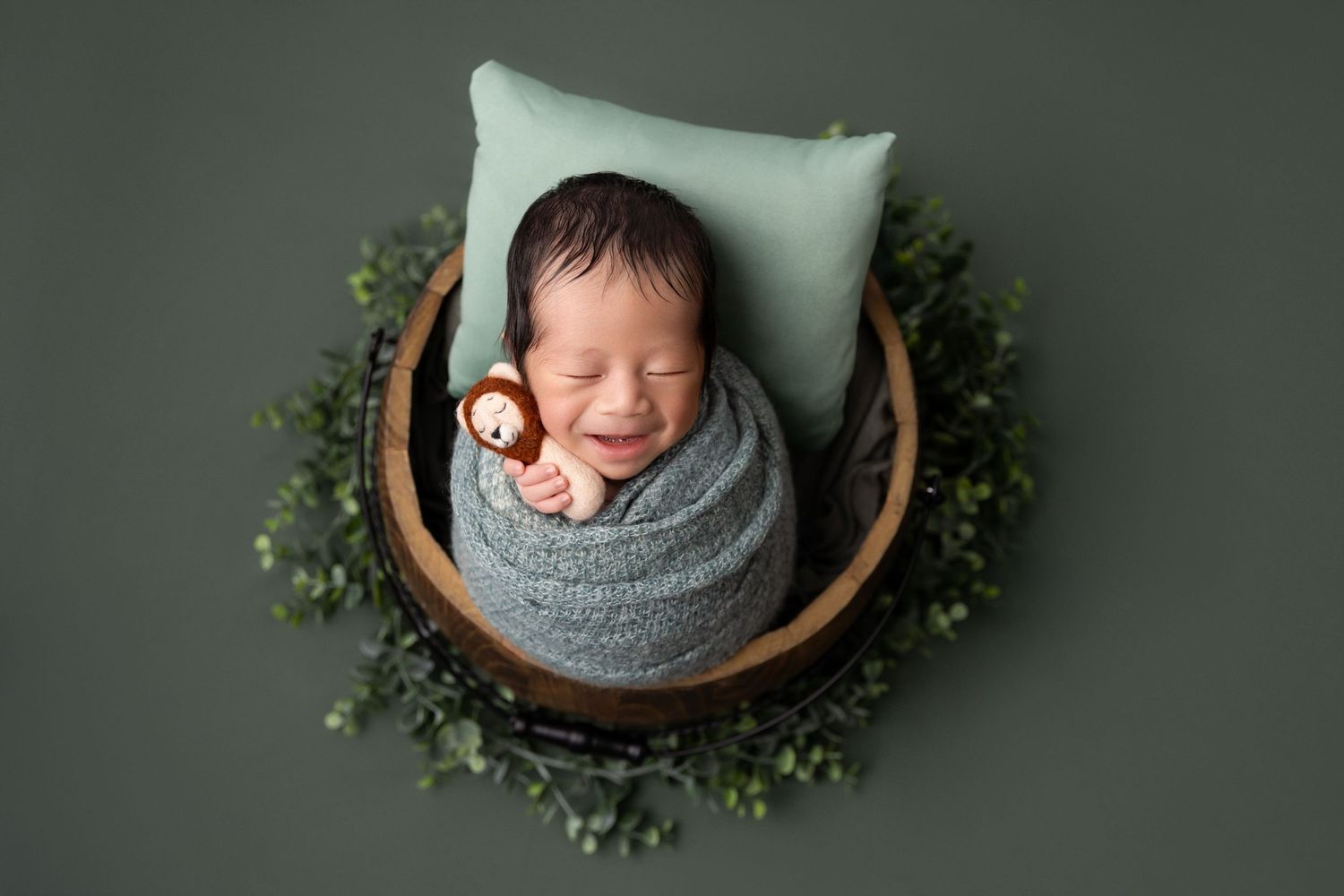 A newborn baby sleeps peacefully in a wooden bowl surrounded by greenery on a dark green backdrop.
