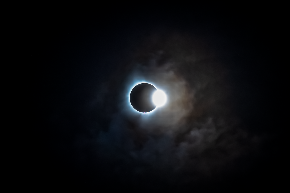 A dramatic solar eclipse with glowing ring of light around the dark moon, surrounded by wispy clouds in dark sky.