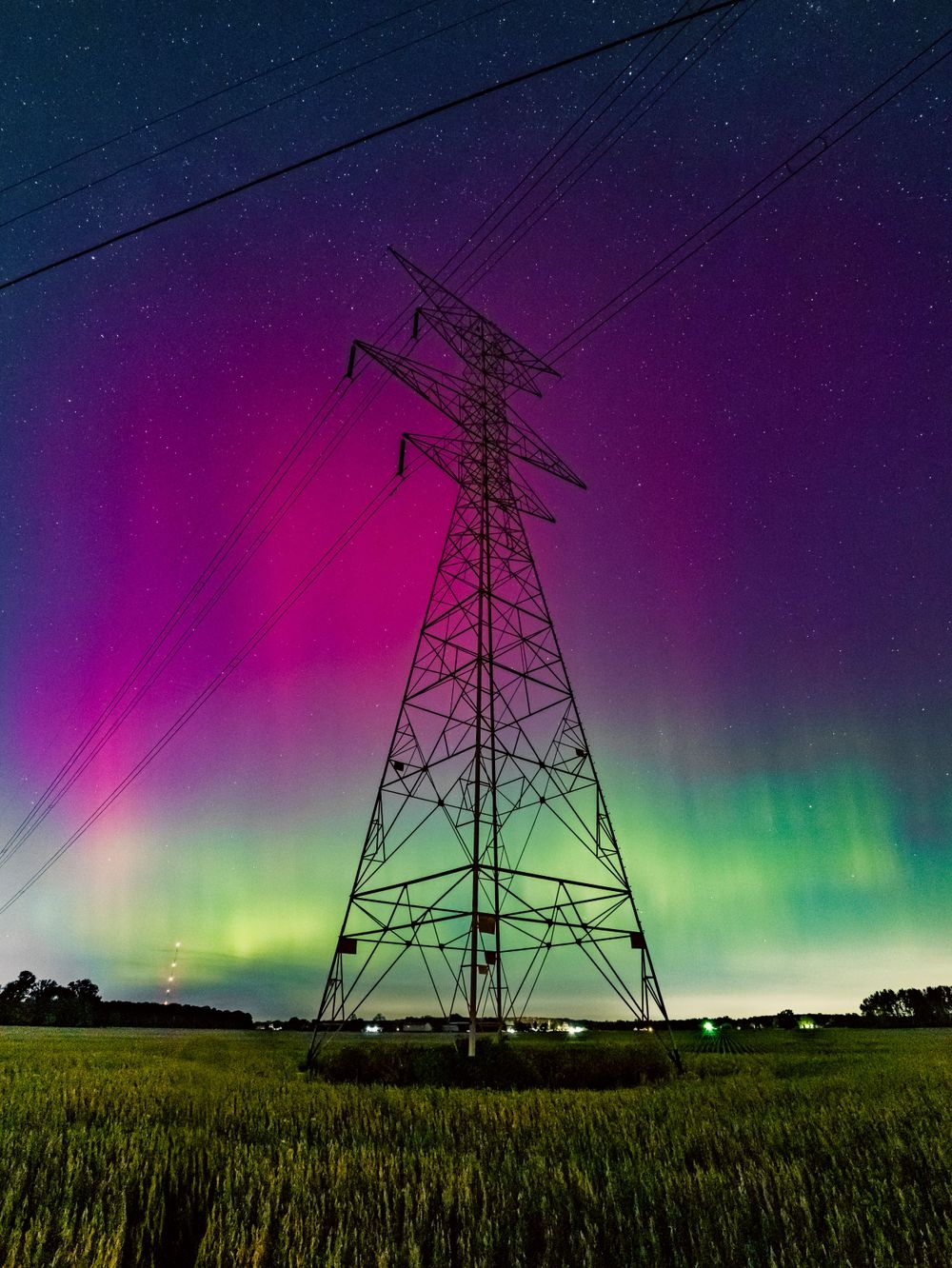 Electric transmission tower silhouetted against vibrant purple and green aurora borealis in the night sky over a grassy field.