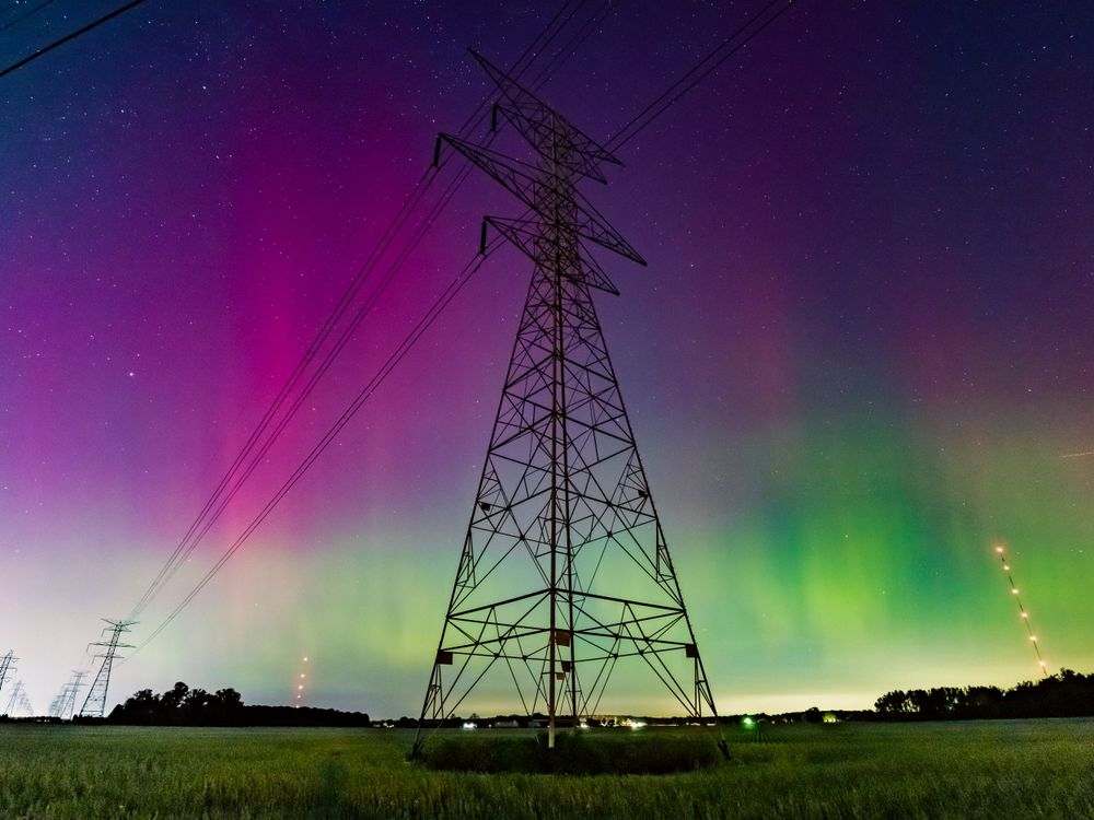 Electrical power transmission tower silhouetted against vibrant purple and green aurora borealis in the night sky.