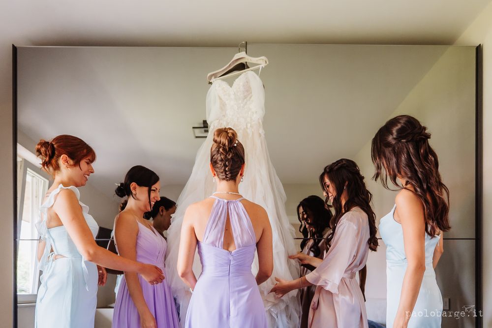 Bridesmaids in pastel purple dresses admiring a white wedding gown hanging in a room.