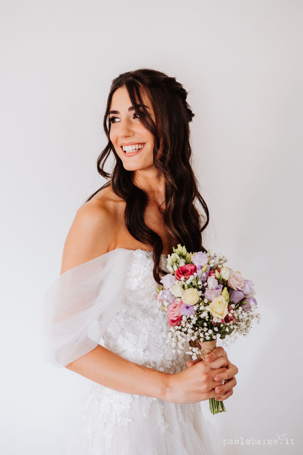 Bride in off-shoulder wedding dress holding colorful bouquet against white background.