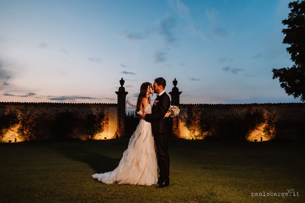 Evening wedding portrait with couple silhouetted against illuminated stone wall at dusk.