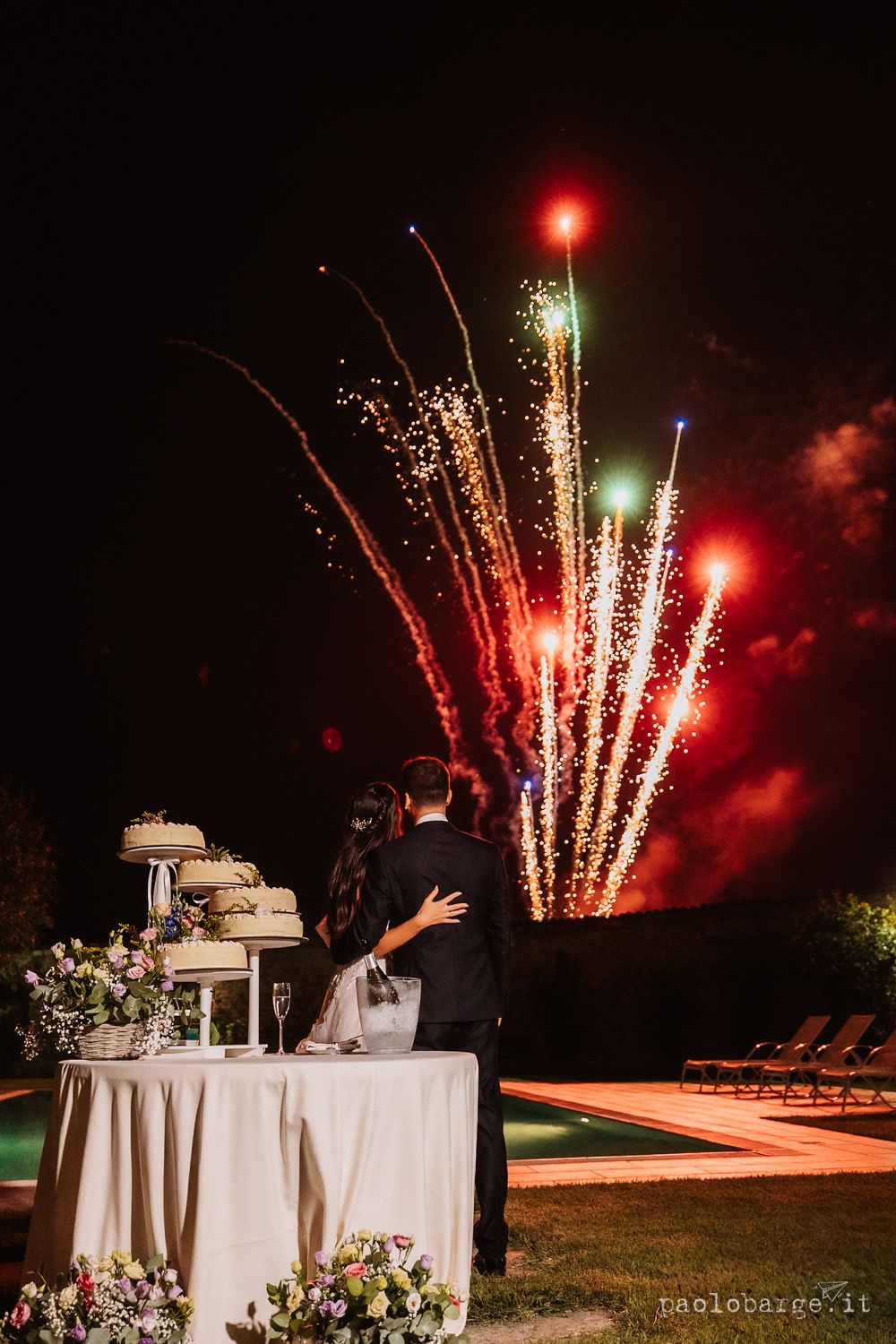 Spectacular fireworks display illuminating the night sky behind a decorated wedding cake table.