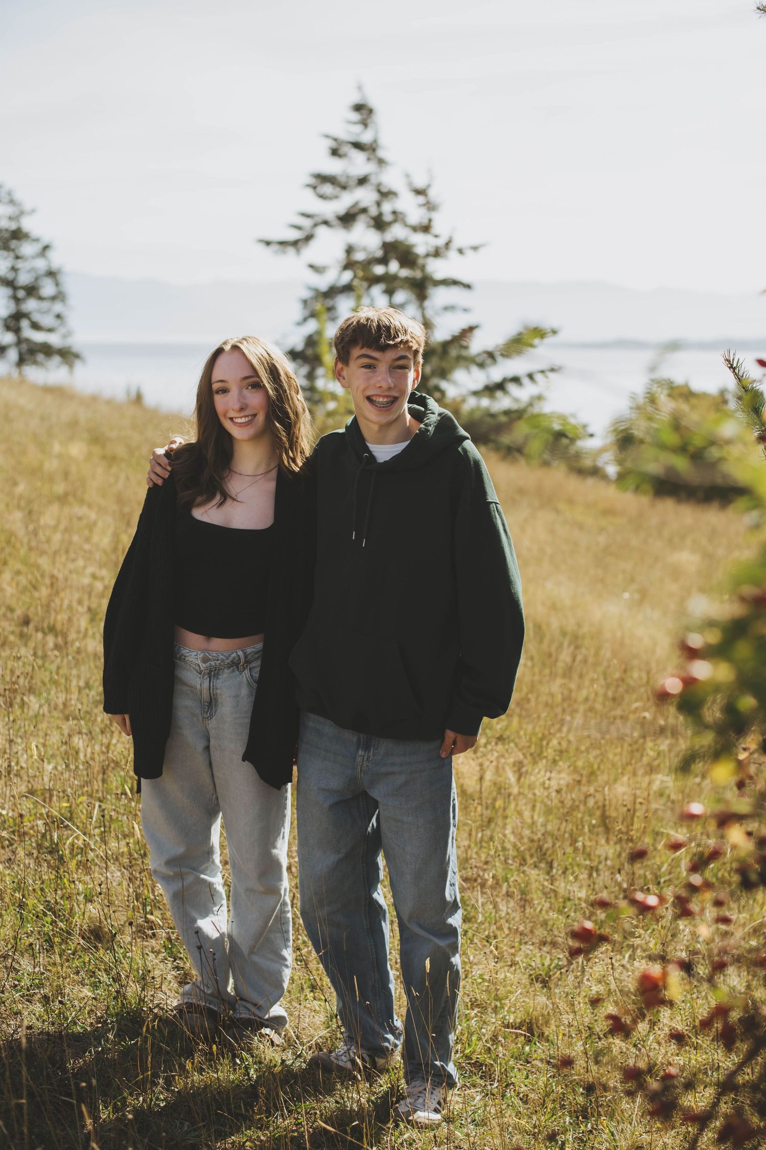 A brother and sister stand close together, smiling in the sunlight, with a grassy field and the ocean in the background.