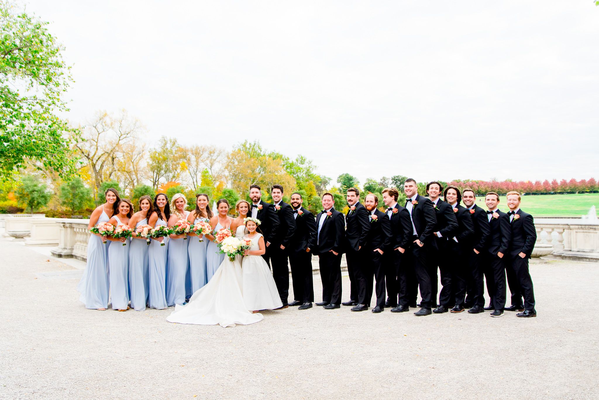 large wedding party in black suits and light blue dresses surround bride and groom at Forest Park in St. Louis