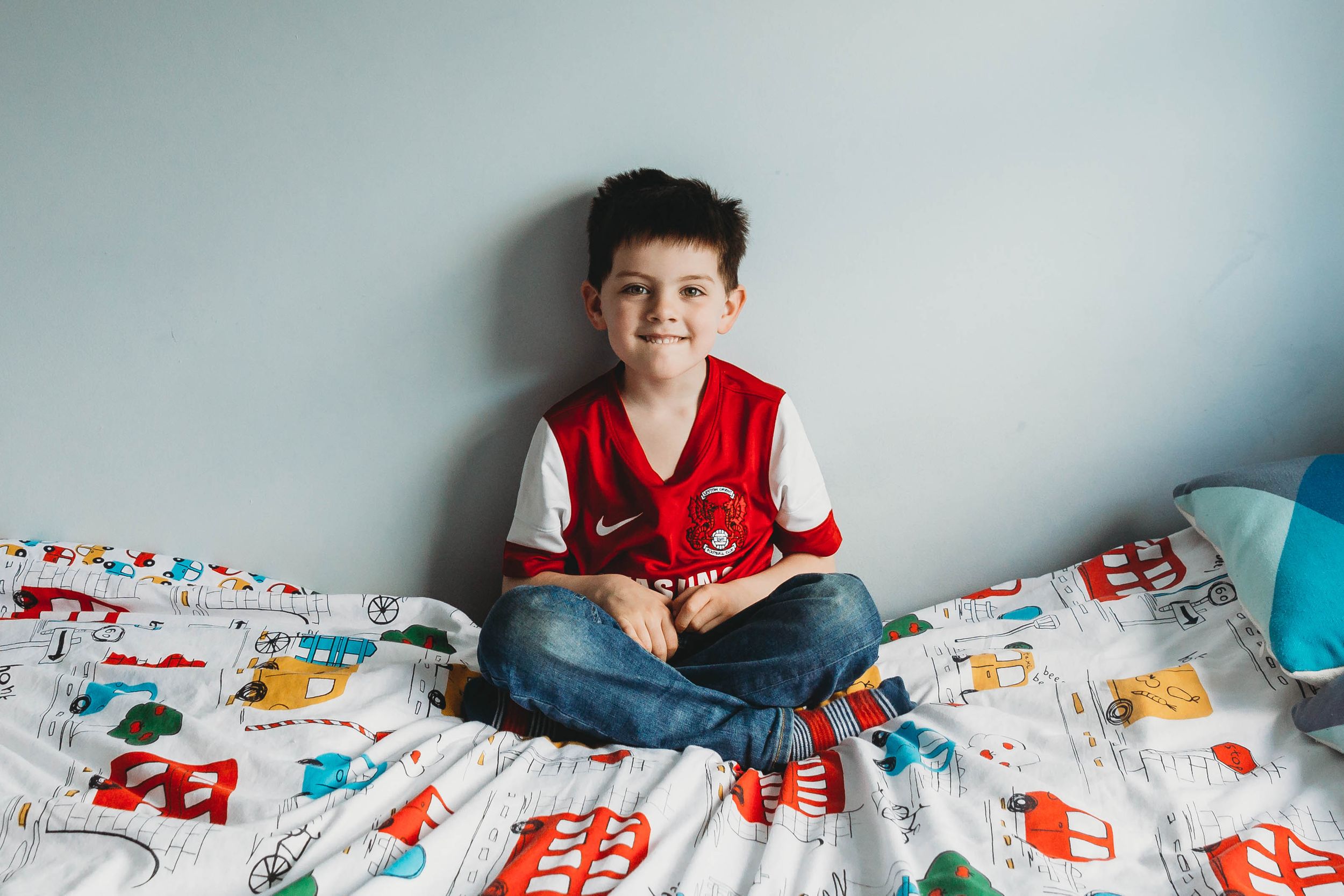 five years old boy sitting on bed wearing leyton orient football shirt grinning at camera