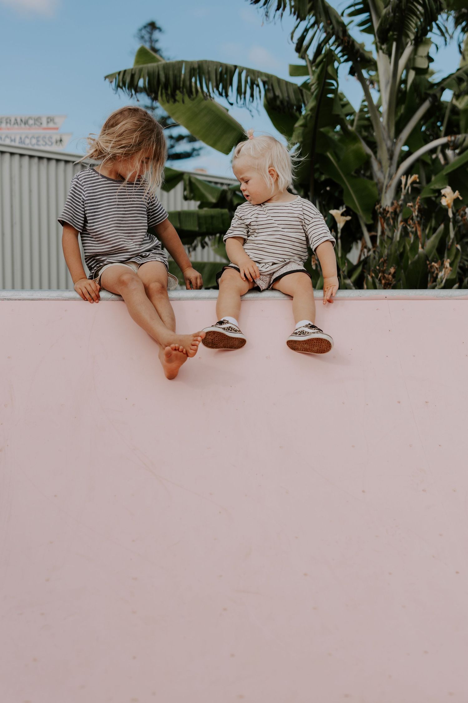 Two children in striped shirts sitting on pink wall with tropical plants in background.