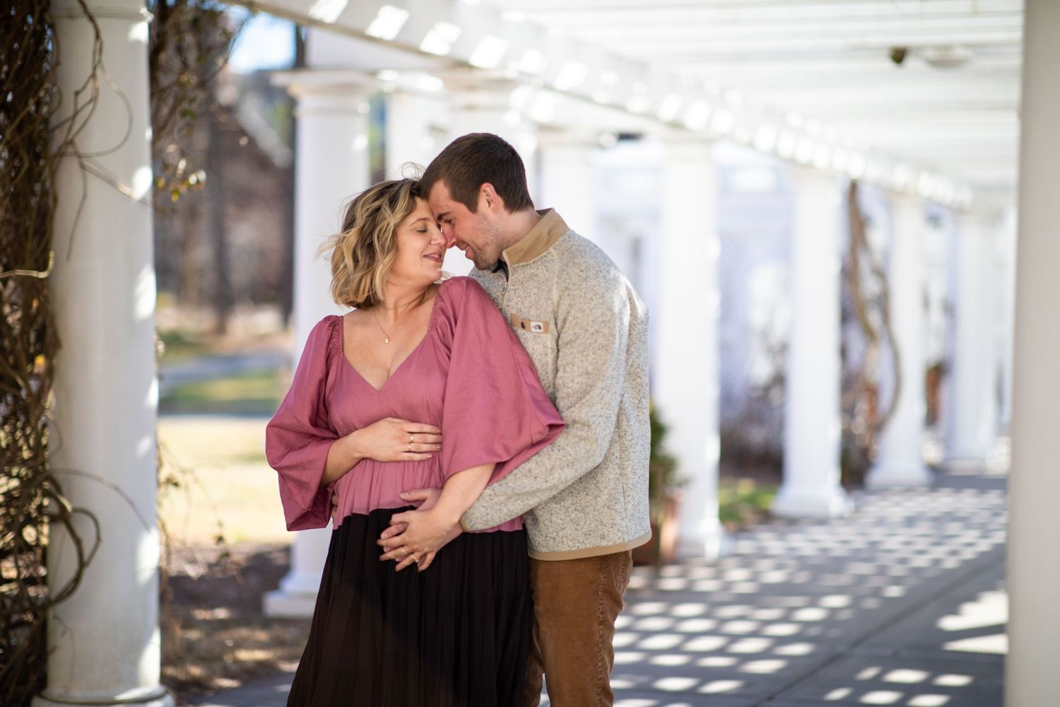 Couple embracing under white columns with dappled sunlight.