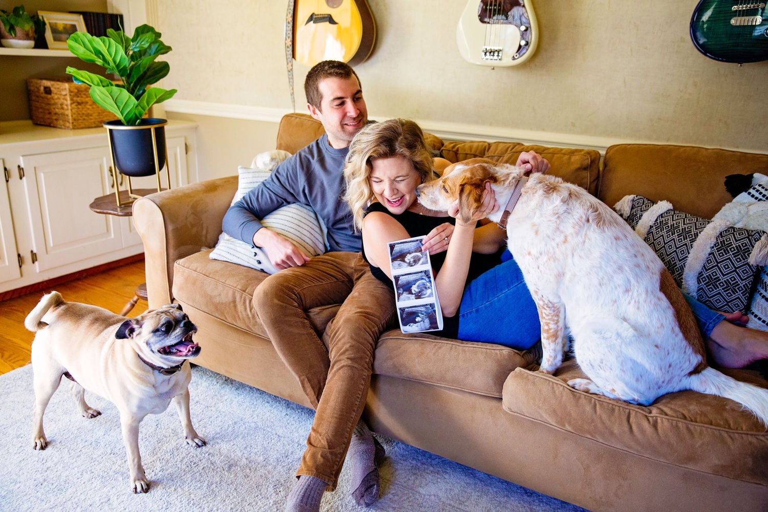 Family relaxing on brown couch with dog in cozy living room decorated with guitars on wall.