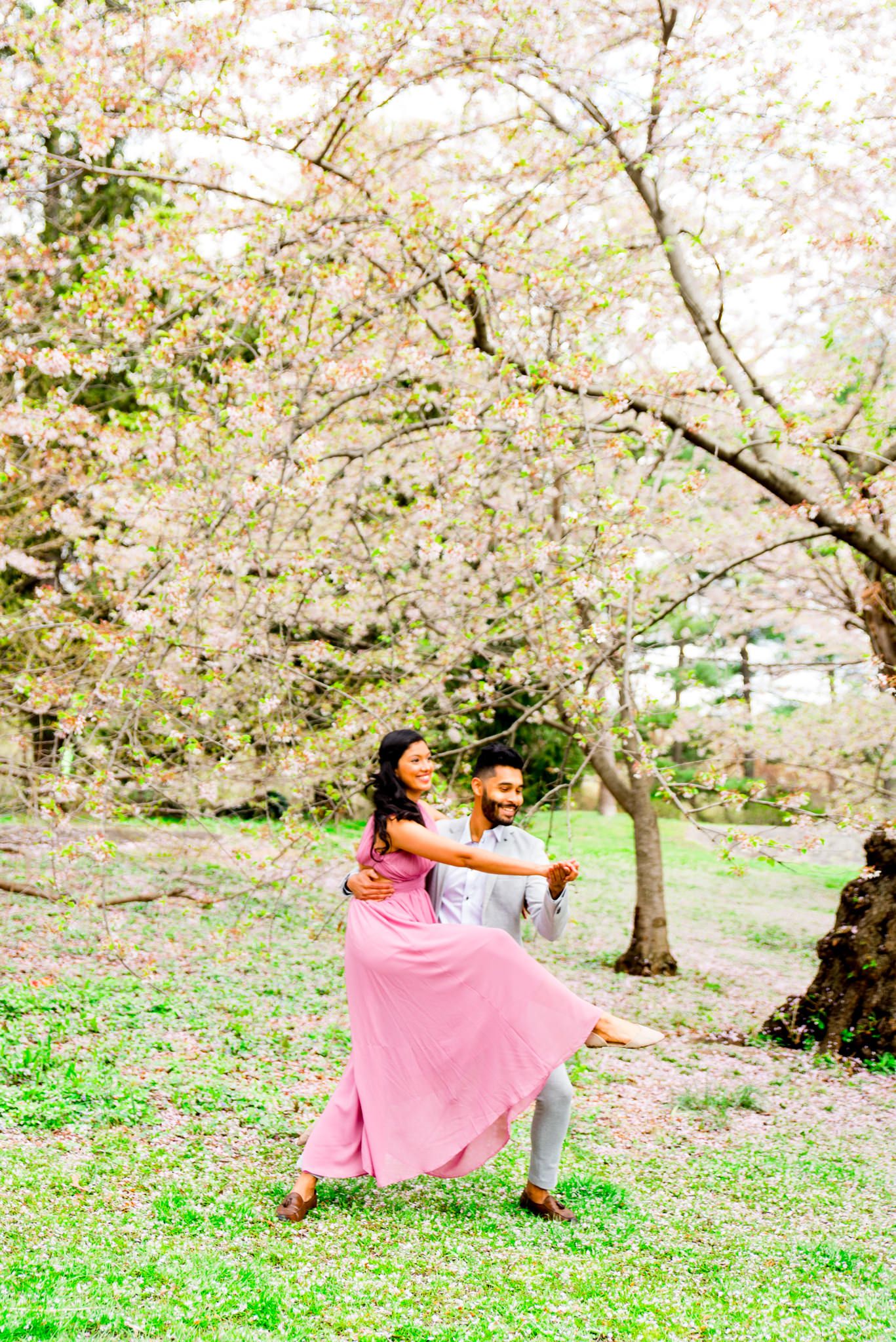 Woman in pink dress and man in gray suit dancing under Central Park Cherry Blossom engagement pictures