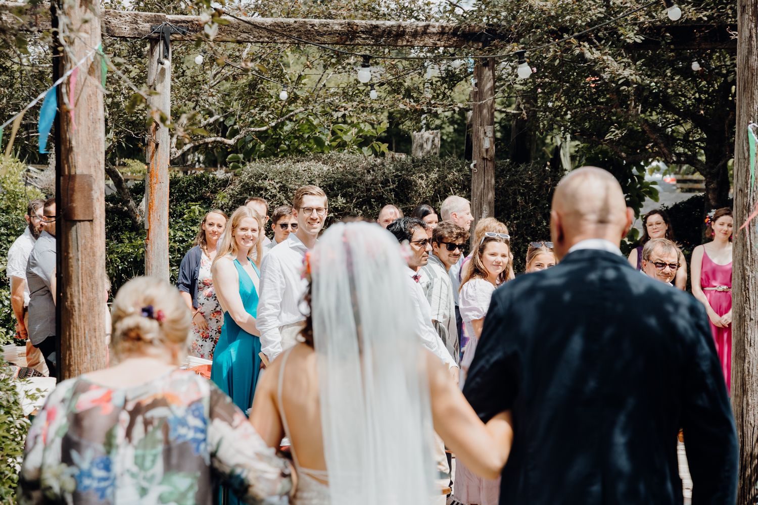 Outdoor wedding ceremony with guests gathered under trees and colorful ribbon decorations.