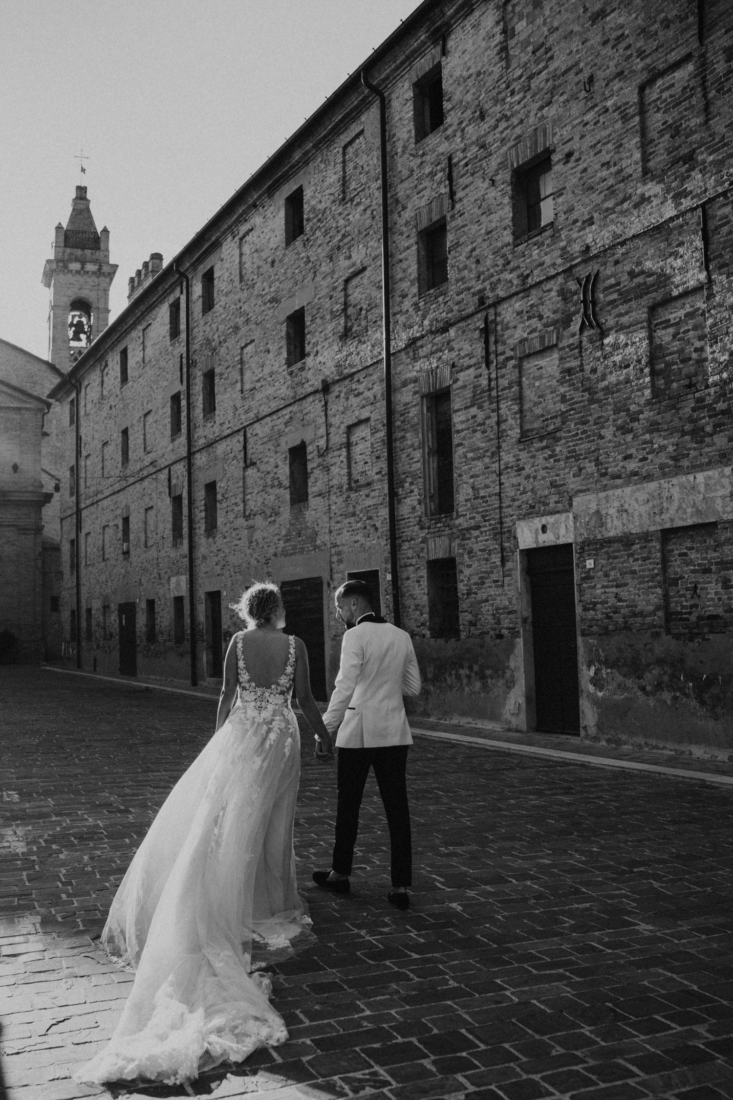 A couple in wedding attire walks along a cobblestone street beside an old stone building.